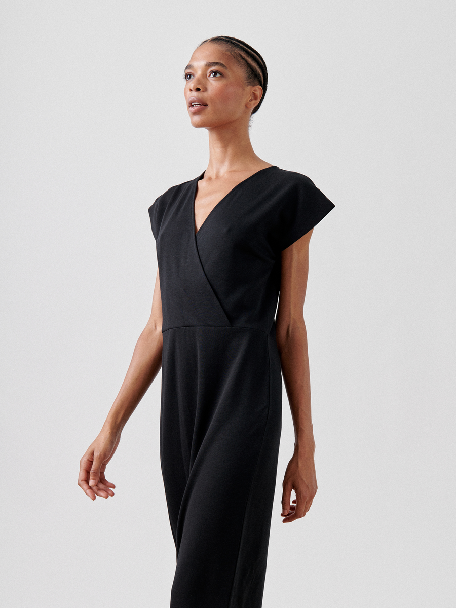 A person with braided hair is wearing a sleeveless, black Wrap Mido Dress by Zero + Maria Cornejo with a V-neckline. They are looking upwards with an expression of confidence and poise. This chic ensemble, reminiscent of a v-neck faux wrap design, stands out against the plain white background.