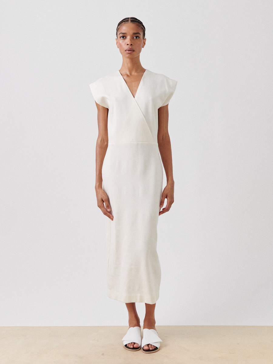 A person stands against a plain white background wearing a sleeveless, V-neck faux wrap beige Wrap Mido Dress by Zero + Maria Cornejo made in New York. The cotton-modal jersey dress boasts a simple, elegant design with a wrap-style front. They are also wearing white slide sandals and have their hair styled in a neat, low bun.