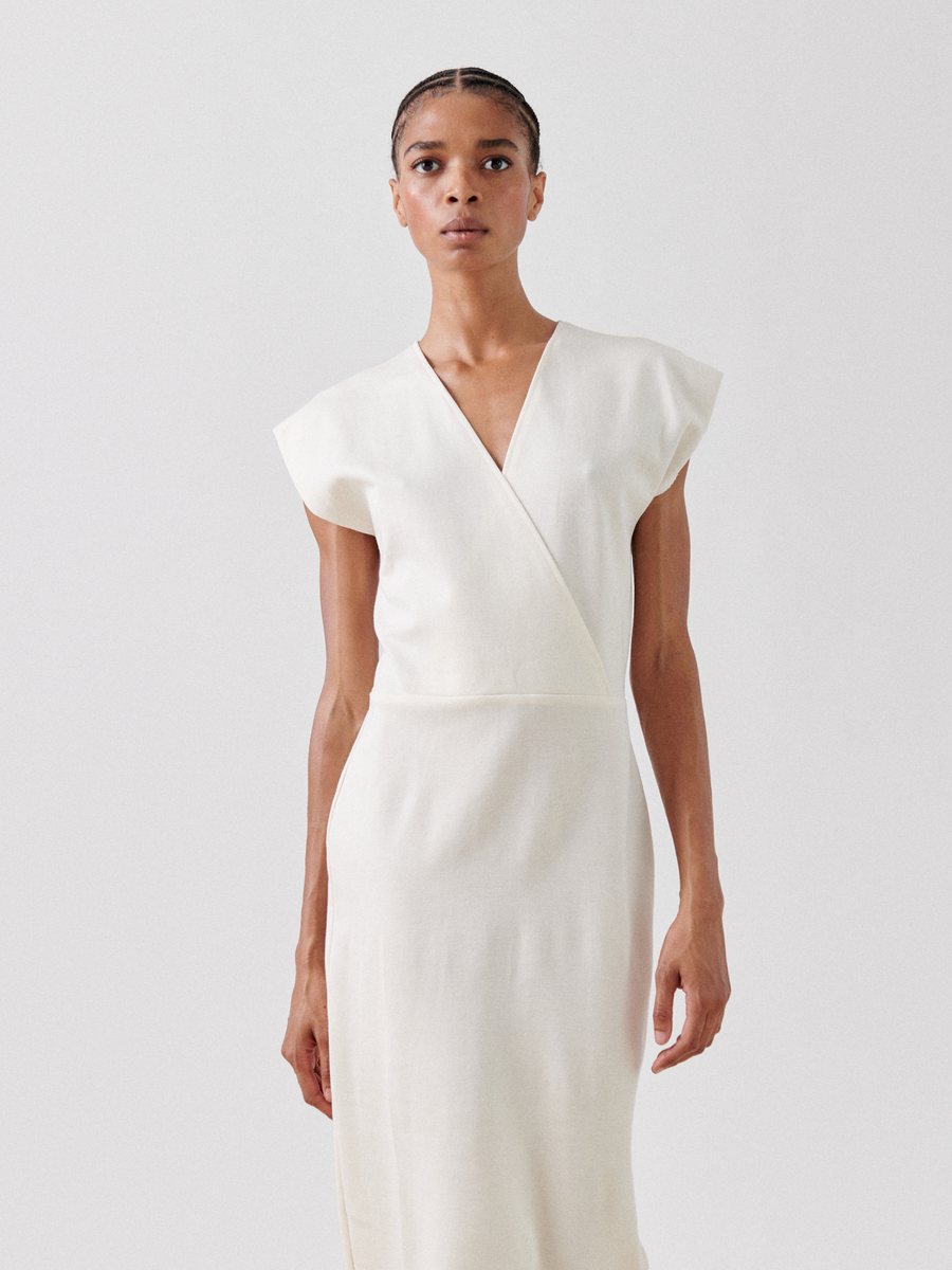 A person with a focused expression is shown from the knees up, standing against a plain white background. They are wearing a sleeveless, V-neck faux wrap, cream-colored Wrap Mido Dress by Zero + Maria Cornejo that fits snugly at the waist and falls straight down. Made in New York from soft cotton-modal jersey, their hair is pulled back as they look directly ahead.