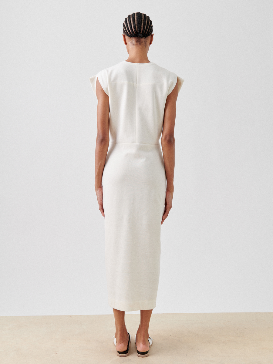 A person with braided hair is standing facing away from the camera, showcasing a sleeveless, off-white, knee-length Wrap Mido Dress by Zero + Maria Cornejo and flat sandals. The background is plain and neutral.