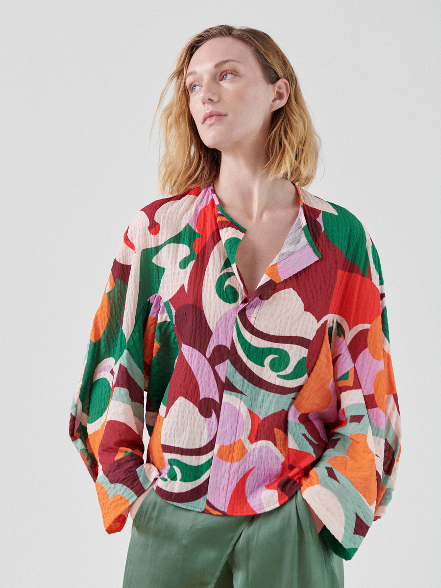 A person with light brown hair wears a brightly colored, multicolor print Long-Sleeved Akeo Shirt by Zero + Maria Cornejo with abstract designs in red, green, orange, purple, and white. They have their hands in the pockets of their sage green pants and are looking slightly to the left. The background is plain and light gray.
