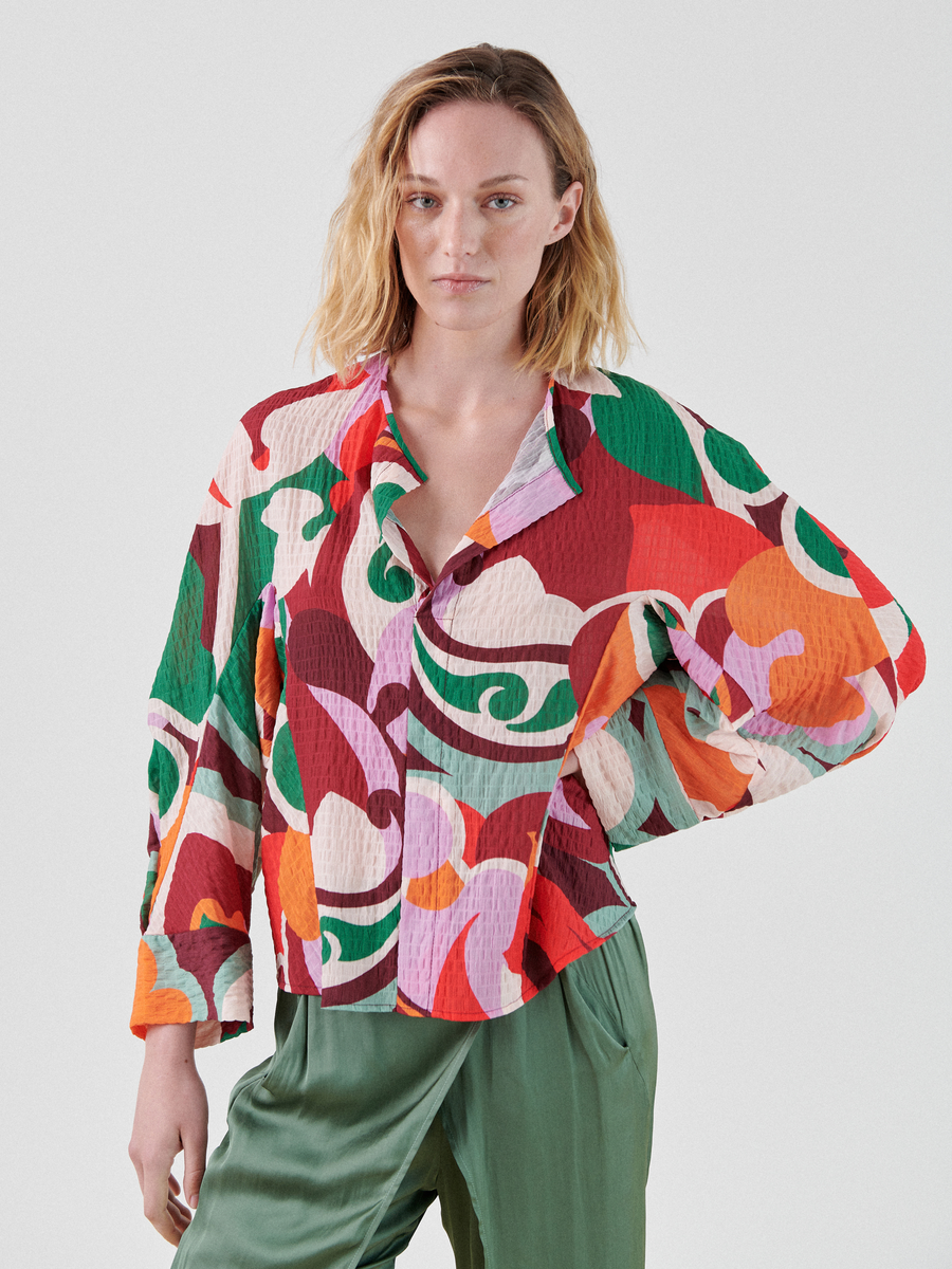 A person with shoulder-length blond hair stands confidently in a Long-Sleeved Akeo Shirt from Zero + Maria Cornejo. They are wearing green pants and looking directly at the camera against a plain background, perfectly dressed for summer occasions.