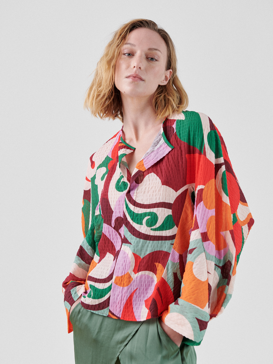 A person with shoulder-length blonde hair is wearing a vibrant, multicolor print Long-Sleeved Akeo Shirt by Zero + Maria Cornejo featuring shades of red, green, pink, and orange. They have their hands in the pockets of their green pants and are standing against a plain, light-colored background. Perfect for summer occasions!