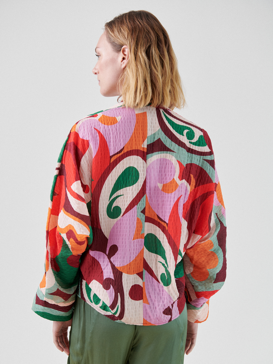 A person with shoulder-length blonde hair is turned slightly away from the camera, wearing a loose-fitting, multicolor print Long-Sleeved Akeo Shirt with shades of red, purple, orange, green, and white by Zero + Maria Cornejo, paired with light green pants. This stylish outfit is perfect for summer occasions and is Made in New York.