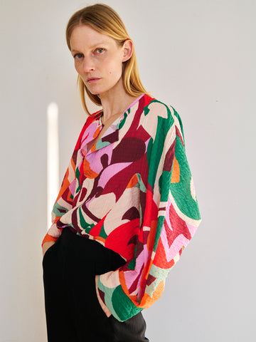 A woman with long blonde hair, wearing a colorful abstract patterned blouse and black pants, stands against a plain background with a neutral expression. The Long-Sleeved Akeo Shirt by Zero + Maria Cornejo features vibrant reds, greens, pinks, and oranges—perfect for summer occasions.