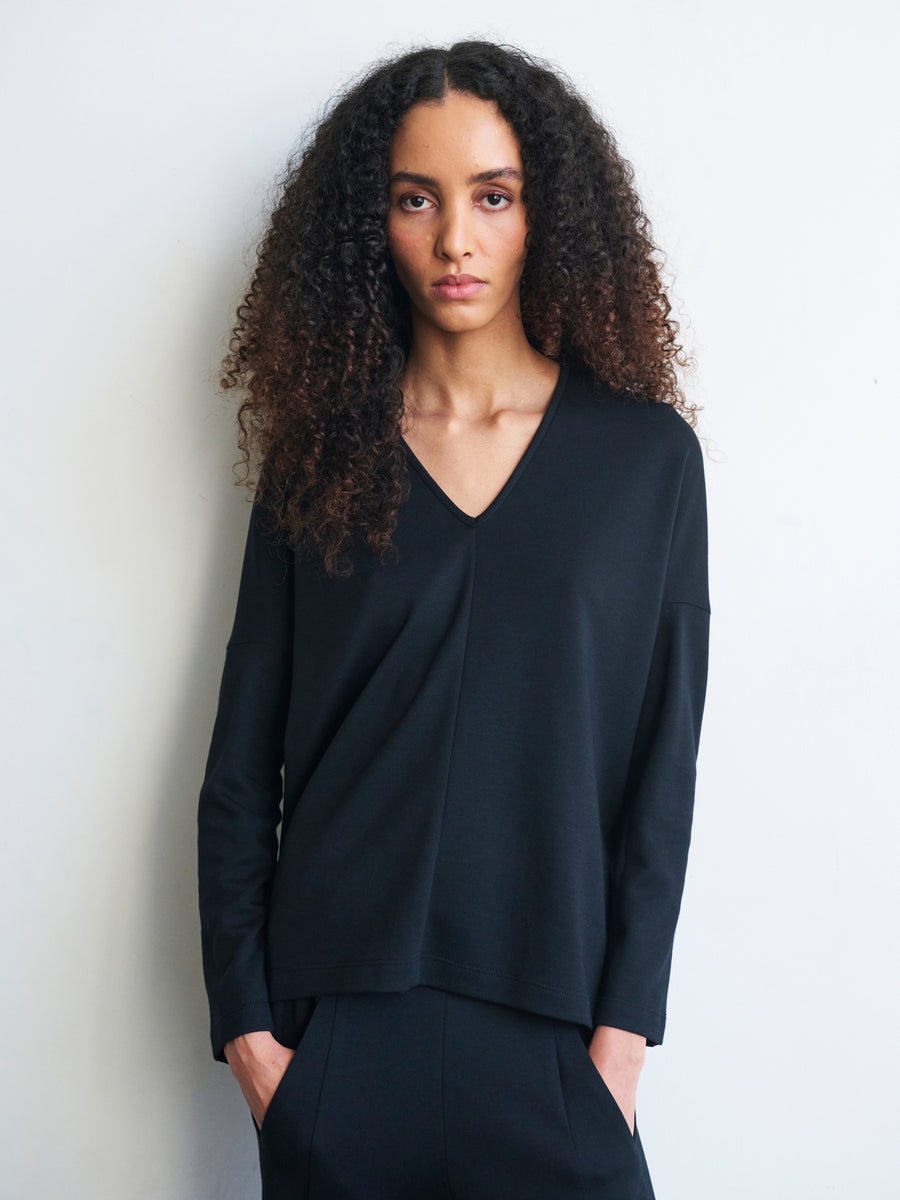 A person with long curly hair wearing a Long-Sleeved Koya Top by Zero + Maria Cornejo, crafted from soft cotton-modal jersey in black, stands against a plain white background, looking at the camera with a neutral expression.