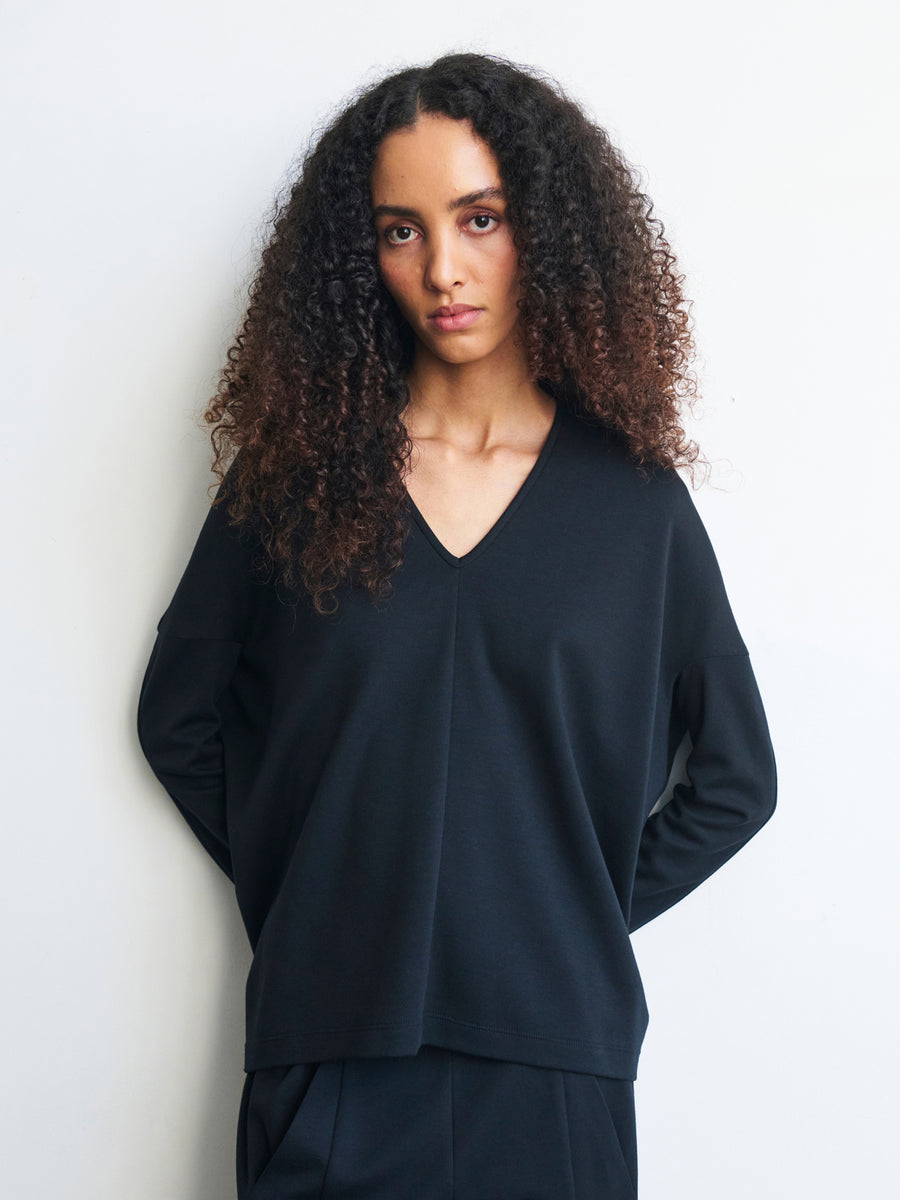 Against a plain background, a person with long, curly hair poses wearing the Long-Sleeved Koya Top from Zero + Maria Cornejo. The loose-fitting cocoon-like silhouette, crafted from soft cotton-modal jersey, features a black V-neck that radiates effortless elegance as they place their hands behind their back.