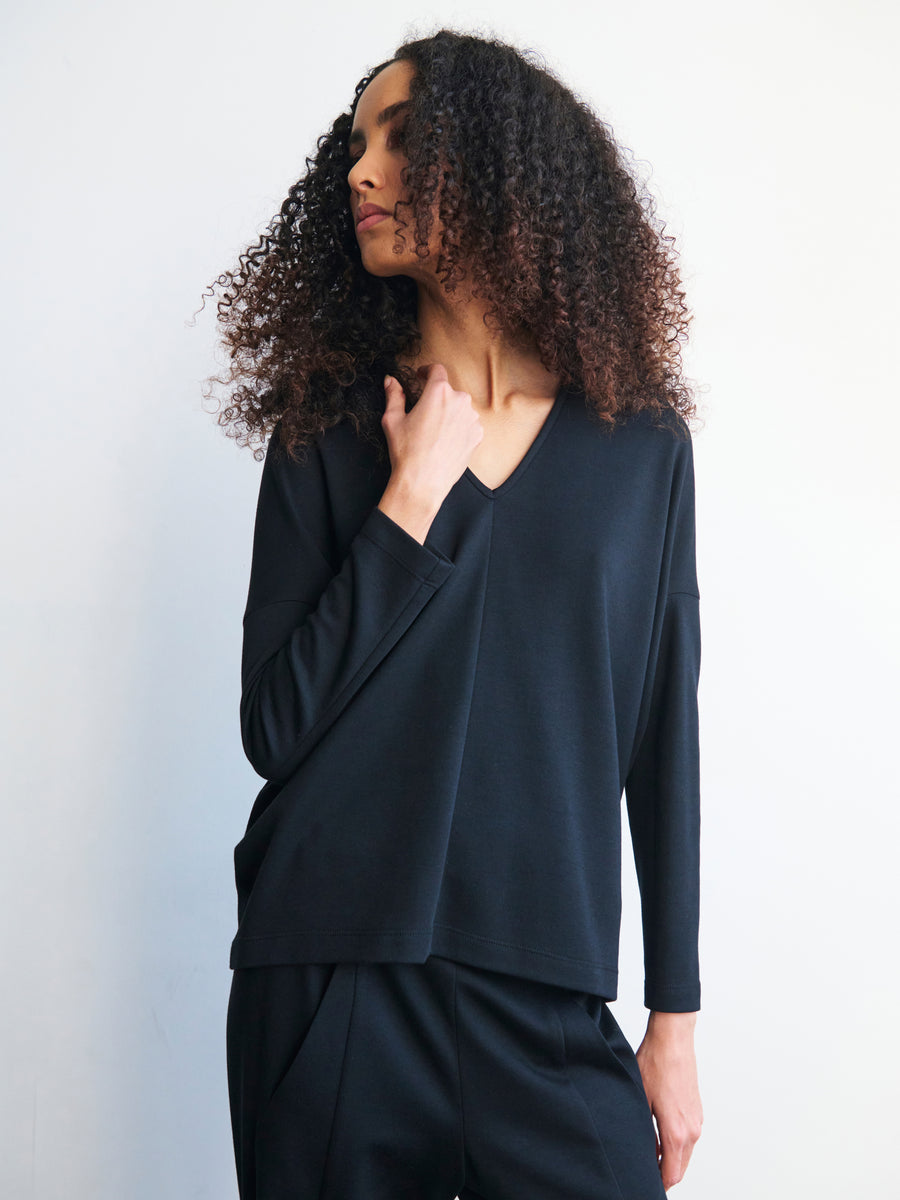 A person with curly hair is wearing a loose-fitting Long-Sleeved Koya Top in black, designed by Zero + Maria Cornejo, along with matching black pants. They are posing against a simple light-colored background in a relaxed posture, with one hand touching their chest.