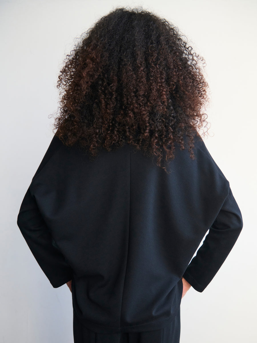 A person with curly hair is wearing the Long-Sleeved Koya Top by Zero + Maria Cornejo in a black cotton-modal jersey fabric, standing facing away against a plain white background.