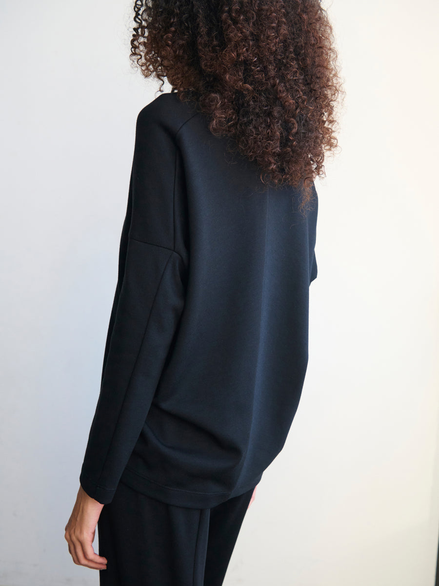 A person with curly hair is facing away from the camera, dressed in a loose-fitting black Long-Sleeved Koya Top by Zero + Maria Cornejo and matching black pants made from a cotton-modal jersey, standing against a plain white background.