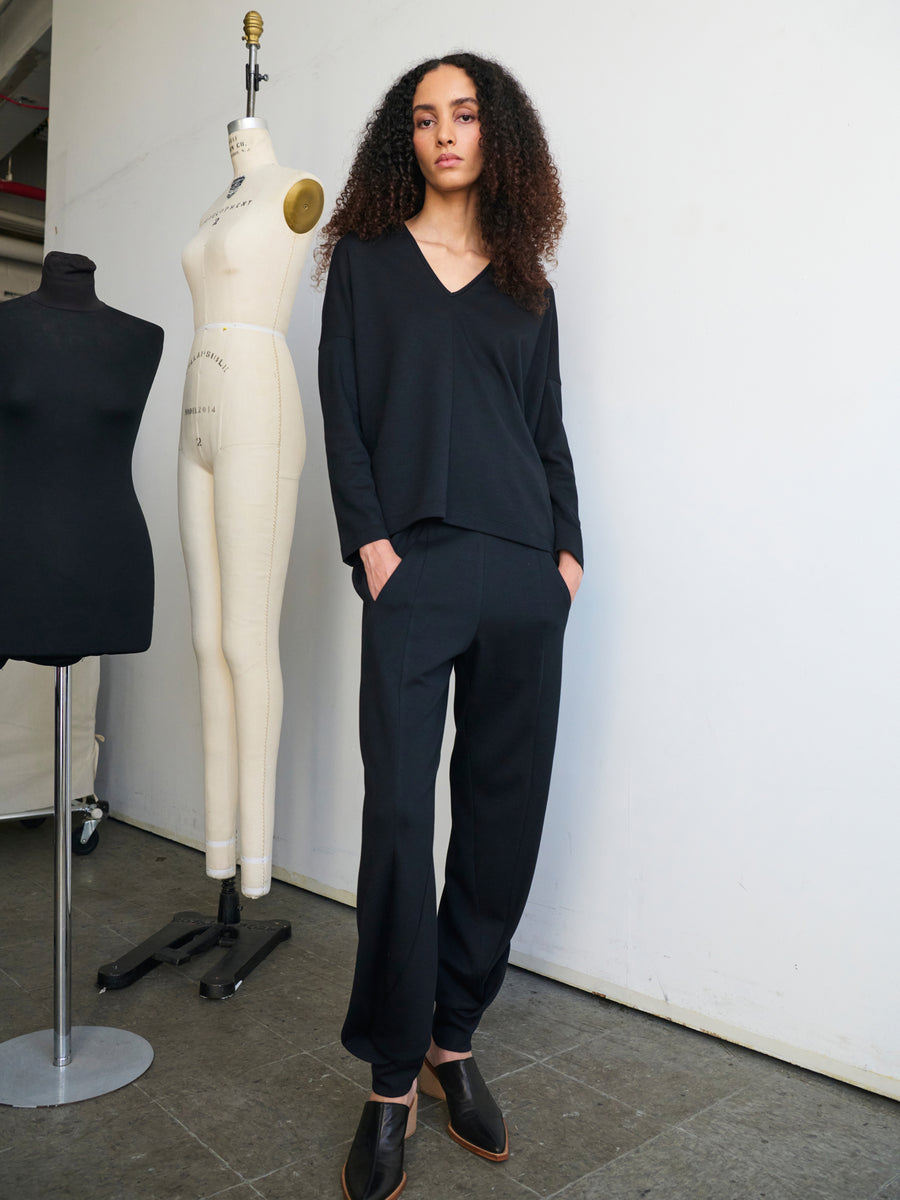 In a fashion studio, an individual with curly hair stands confidently, dressed in a sleek black ensemble featuring the Jersey Cuff Akeo Pant by Zero + Maria Cornejo. The pants have an elastic banded waist and are paired with black shoes for a complete look, while two dress forms stand in the background.