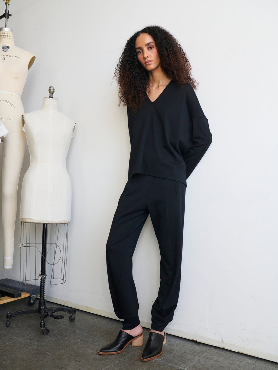 A person with curly hair is leaning against a wall, wearing a black V-neck top and the Jersey Cuff Akeo Pant by Zero + Maria Cornejo. They stand next to two dress forms in a fashion studio with a minimalist aesthetic.