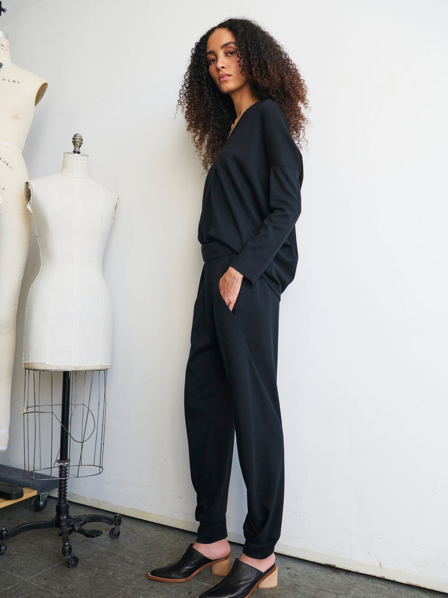 A person with long curly hair stands confidently, wearing the fashionable Jersey Cuff Akeo Pant by Zero + Maria Cornejo paired with brown shoes. Beside them, two dress forms display designs made from sustainable materials in a well-lit room, exuding a fashion-forward and creative atmosphere.