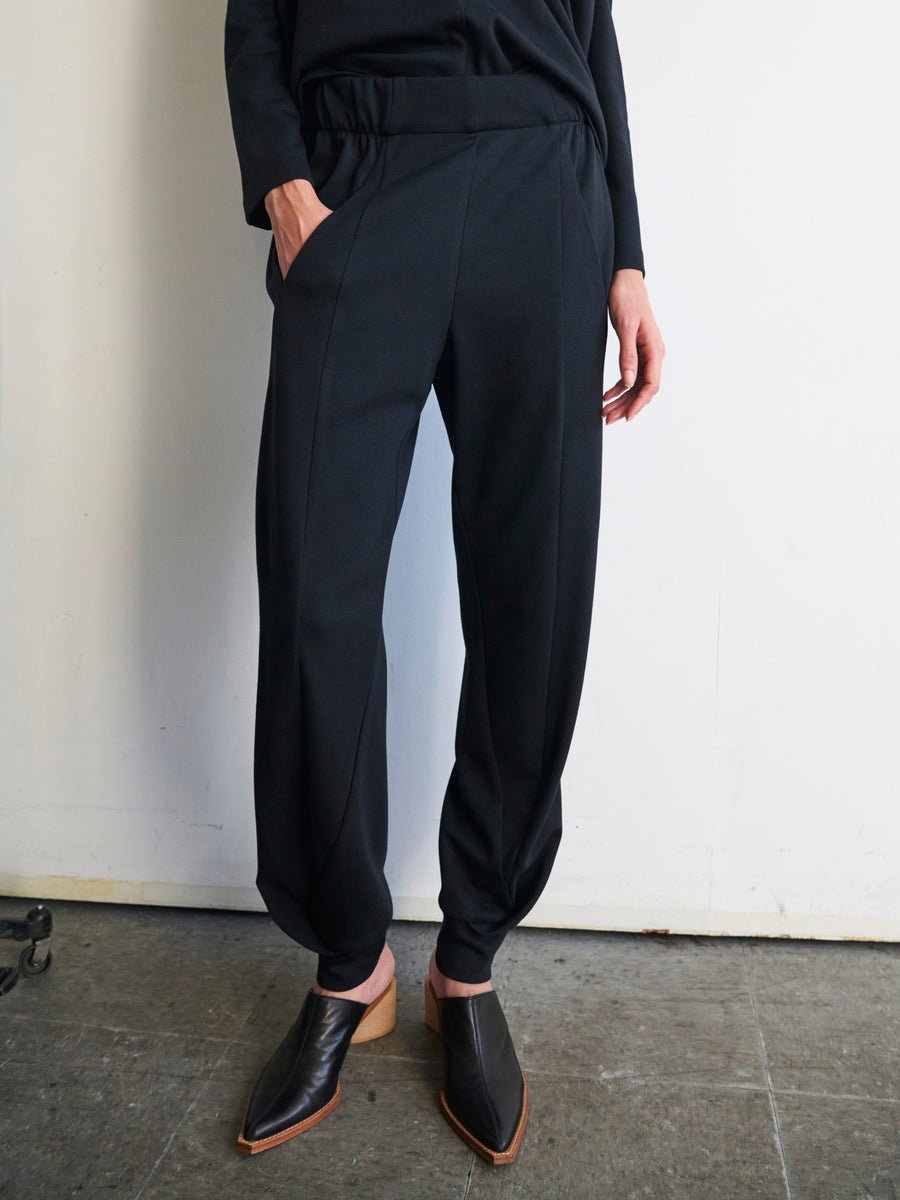 A person wearing the Jersey Cuff Akeo Pant by Zero + Maria Cornejo, featuring a sleek cotton-modal jersey fabric and an elastic banded waist, along with black pointed shoes, stands against a white wall, one hand casually in their pocket. The use of sustainable materials beautifully complements the industrial touch of the concrete floor.