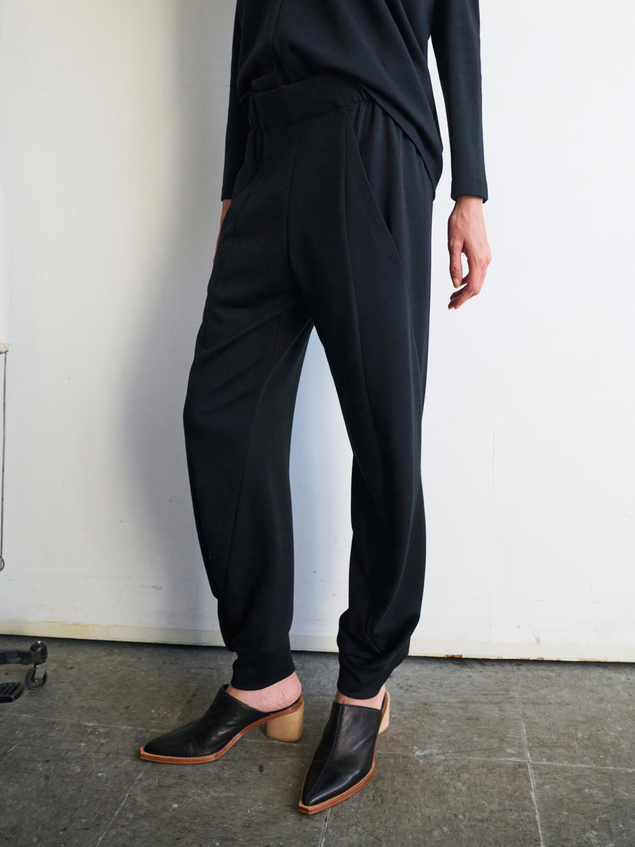 A person stands against a plain white wall, dressed in black Jersey Cuff Akeo Pants by Zero + Maria Cornejo. These loose-fitting cotton-modal jersey pants feature an elastic banded waist and are paired with a matching top. Their brown heeled slip-on shoes create an elegant contrast with the gray textured floor, highlighting sustainable materials through style.