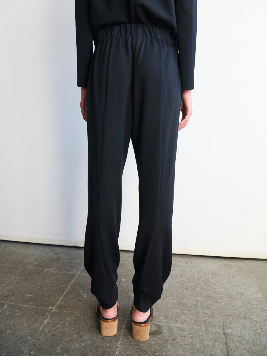 The image features a person standing with their back to the camera, wearing black Jersey Cuff Akeo Pants by Zero + Maria Cornejo and a black top. The loose-fitting pants, made from bio-based materials, offer an effortlessly stylish appearance. Block-heeled sandals finalize the look against a plain white wall backdrop.