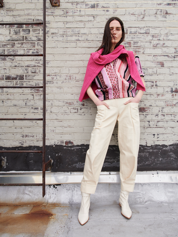 A person stands against a brick wall, wearing a patterned top and Cradle 2 Cradle Certified light organic denim pants. The look is completed with Zero + Maria Cornejo's Akeo Jodhpur ankle boots and a pink sweater draped over their shoulders. A metal ladder is visible on the left.