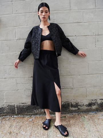 A woman stands against a beige brick wall. She wears a black crop top, black jacket with a subtle pattern, and a Zero + Maria Cornejo Bias Slip Skirt with a side slit. Her hair is tied back, and she gazes directly at the camera with her arms slightly extended to the sides.