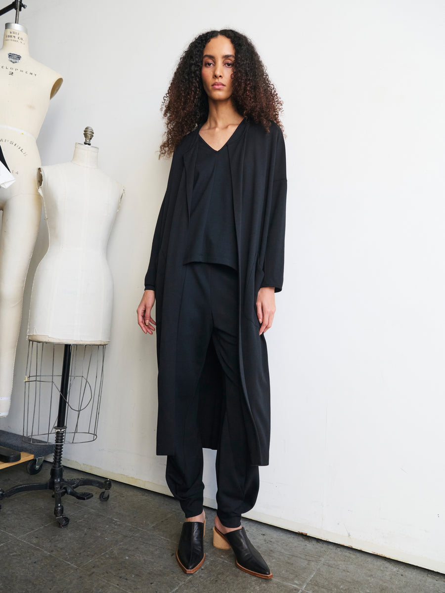 A person with curly hair stands in a minimalist studio, wearing the Jersey Koya Coat by Zero + Maria Cornejo over a black outfit and pointed shoes. Two dress forms are in the background, with one draped in fabric to feature a cocoon silhouette.