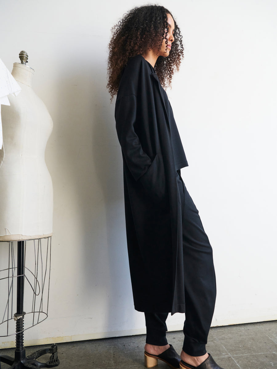 A person with curly hair stands in profile, wearing the Jersey Koya Coat from Zero + Maria Cornejo and black pants, looking at a dress form. They appear in a minimalist studio setting with natural light casting soft shadows.