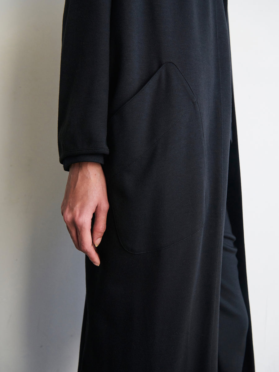 A person is wearing the Jersey Koya Coat by Zero + Maria Cornejo, which is a long, black cotton-modal jersey coat with a draped cocoon silhouette and a prominently visible side pocket. Their hand casually rests along their body, partly covering the pocket against a plain off-white wall in the background.