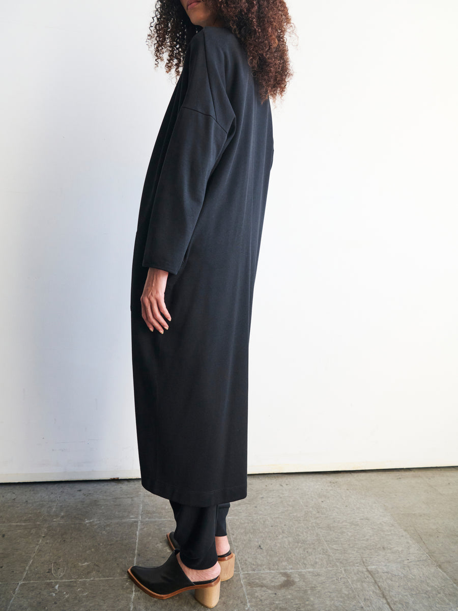 A person with long curly hair stands sideways, wearing the Jersey Koya Coat made of cotton-modal jersey by Zero + Maria Cornejo. They're indoors on a concrete floor against a white wall, their black pants enhancing the softly flowing attire. Their face is not visible.