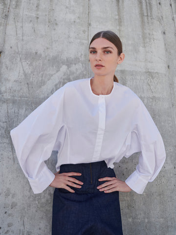 Against a concrete wall, someone stands wearing Zero + Maria Cornejo's Long-Sleeved Akeo Shirt with voluminous sleeves and blue high-waisted pants. The shirt's round neckline complements their composed expression as they place their hands on their hips.