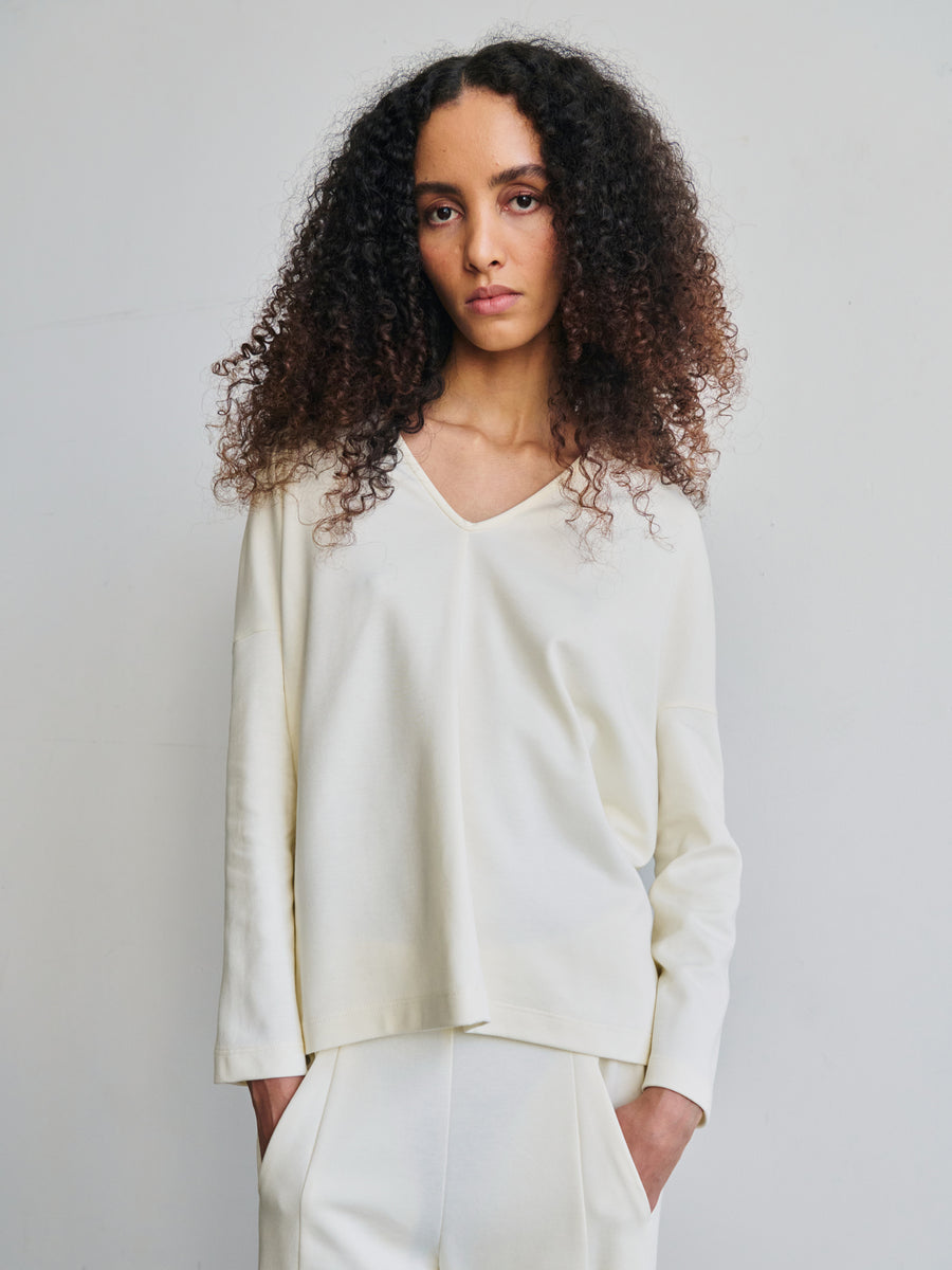 A person with long curly hair is wearing the Long-Sleeved Koya Top from Zero + Maria Cornejo, designed in an off-white V-neck sweater style with a cocoon-like silhouette and matching pants. They stand against a plain background, crafted from soft bio-based materials, exuding a calm expression with hands resting in their pockets.