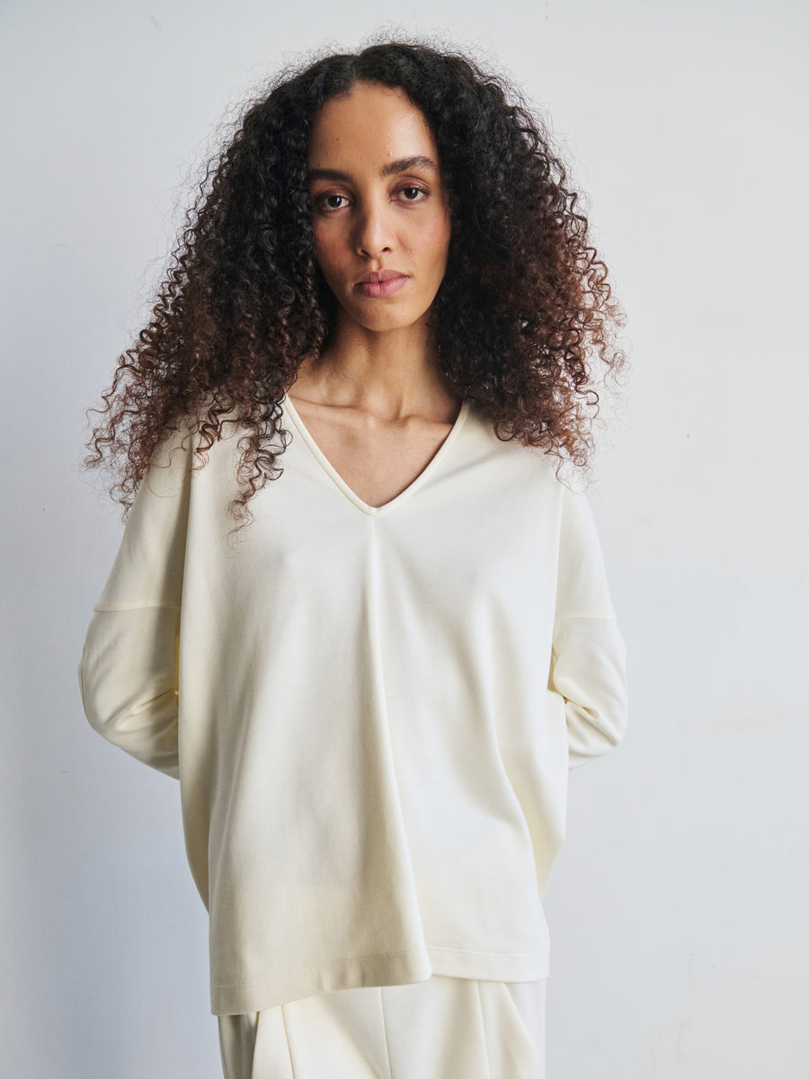 A person with long curly hair is wearing the Long-Sleeved Koya Top by Zero + Maria Cornejo, made from soft cotton-modal jersey in a light cream color. They are standing against a plain background with hands behind their back, maintaining a neutral expression in what appears to be a well-lit studio.