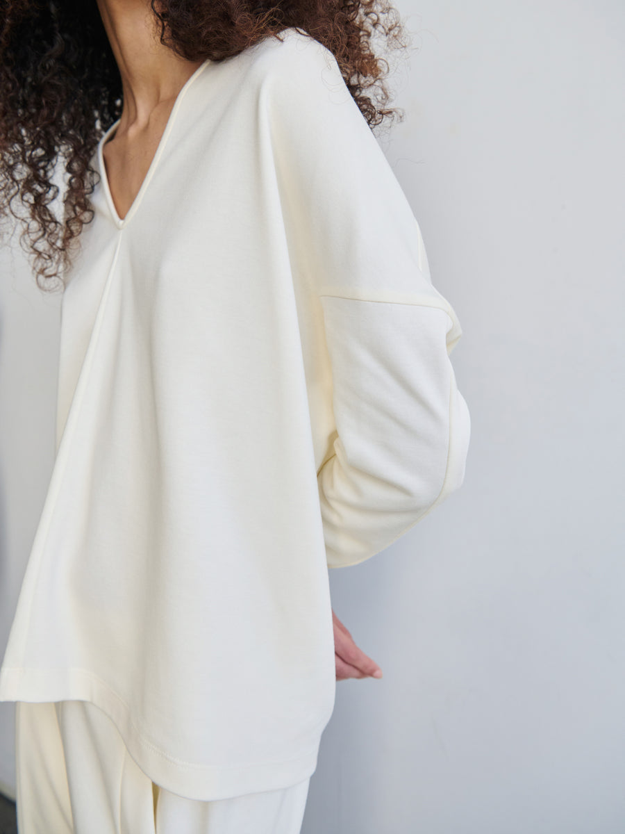 A person with curly hair is wearing an oversized Long-Sleeved Koya Top in light cream, crafted from cotton-modal jersey, along with loose-fitting trousers. They are standing against a plain light background, and the cocoon-like silhouette of the Zero + Maria Cornejo design adds an intriguing touch. The person's face remains unseen.