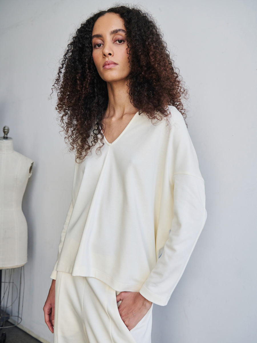 A person with curly hair wearing a loose-fitting Long-Sleeved Koya Top by Zero + Maria Cornejo and pants stands against a plain wall, with a mannequin partially visible in the background.