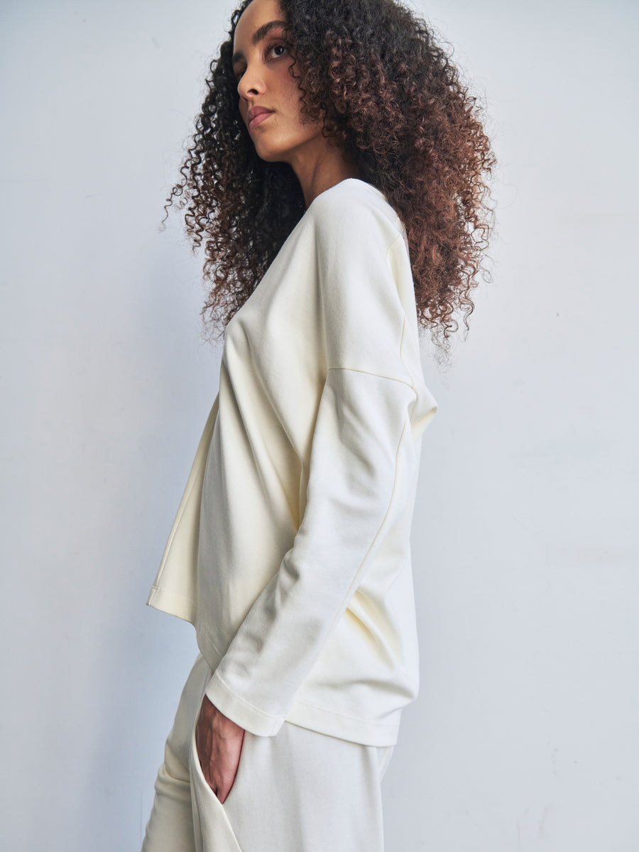 A person with curly hair is dressed in a light-colored Long-Sleeved Koya Top and matching pants from Zero + Maria Cornejo, crafted from cotton-modal jersey. Standing against a plain background, the cocoon-like silhouette of the outfit exudes comfort and casual elegance.