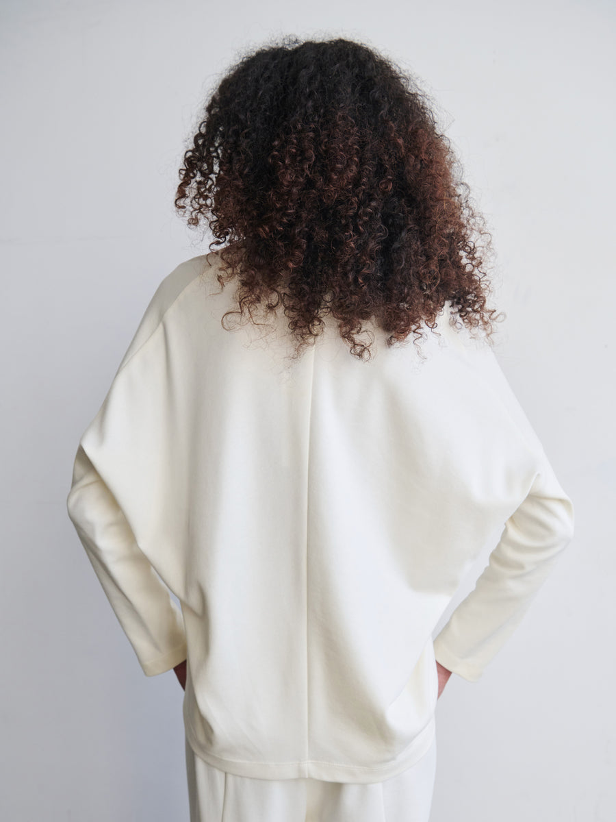 A person with curly hair, draped in the Long-Sleeved Koya Top by Zero + Maria Cornejo, stands facing a white wall. The loose fit of the cream-colored sweater with its cocoon-like silhouette made from cotton-modal jersey enhances their relaxed posture, arms resting gently at their sides.