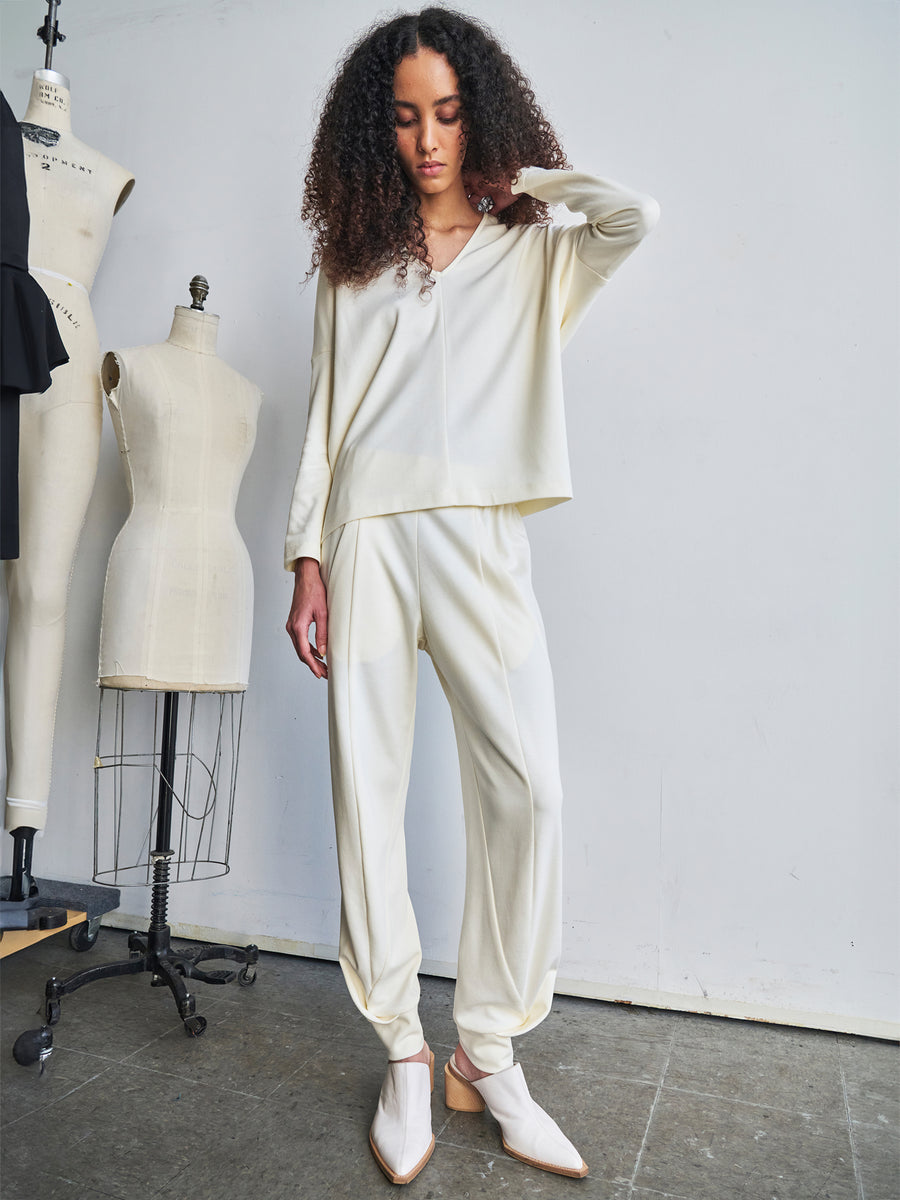 Someone with long, curly hair is wearing the cream-colored Jersey Cuff Akeo Pant by Zero + Maria Cornejo and white pointed shoes. They are standing next to dress forms in a bright room, highlighting sustainable production using bio-based materials.