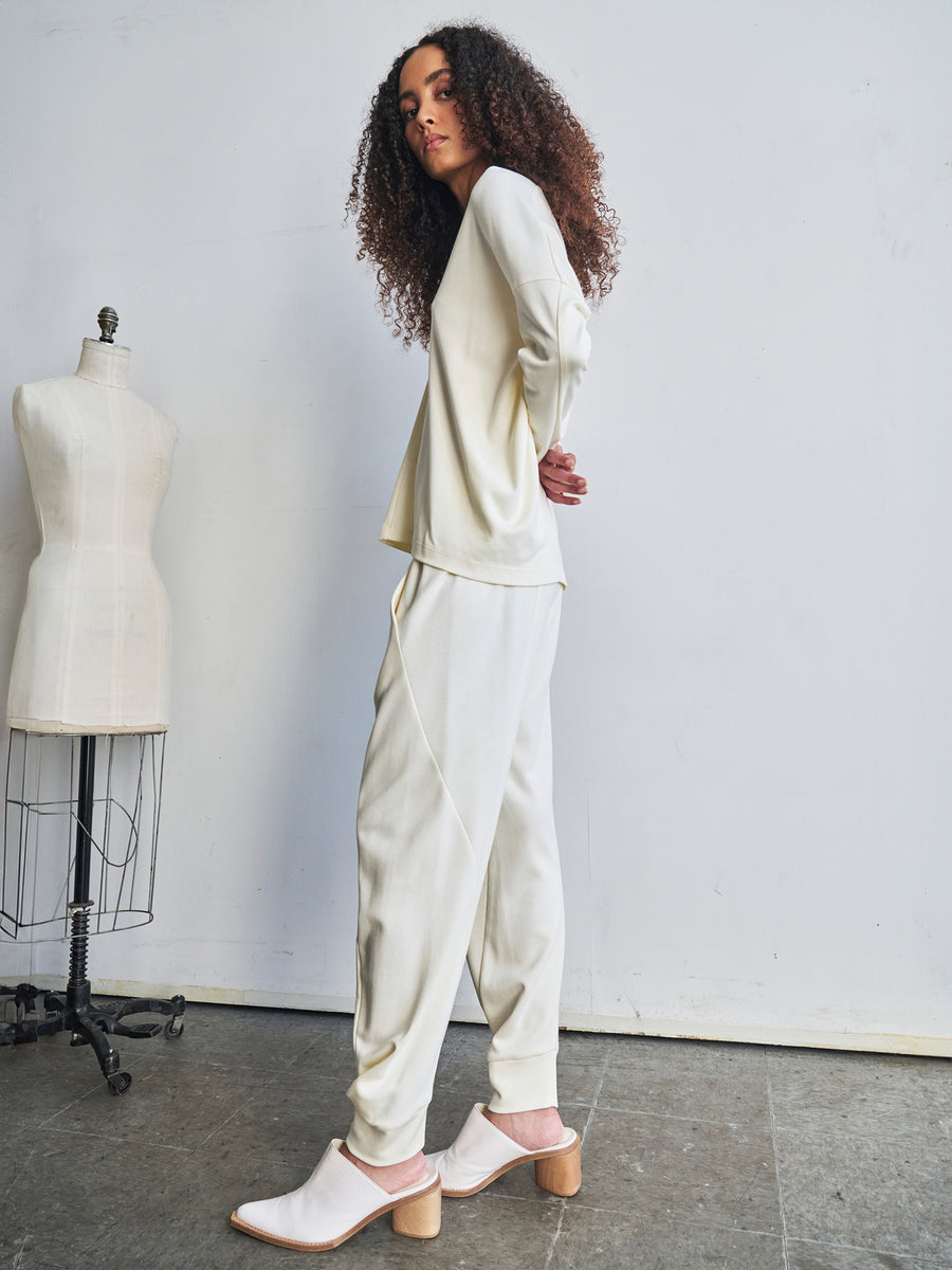 In a minimalist studio setting, a person with curly hair models the cream-colored long-sleeve top paired with the Jersey Cuff Akeo Pant from Zero + Maria Cornejo. Crafted from bio-based materials, they stand sideways, glancing back over their shoulder. In the background, a tailor's mannequin signifies the brand's commitment to sustainable production.