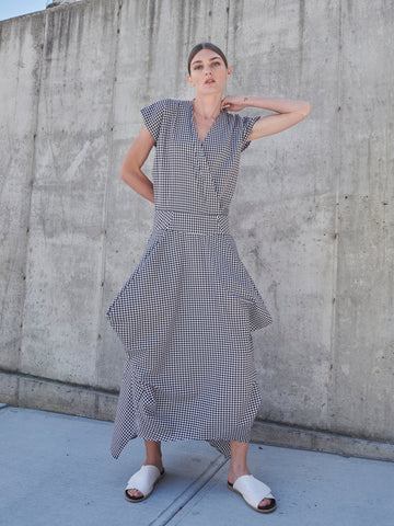 A woman stands against a concrete wall wearing the Wrap Nio Dress by Zero + Maria Cornejo, featuring cap sleeves and an asymmetrical hem in a houndstooth-like checkered pattern. Her hair is pulled back, and she completes the look with white slide sandals.