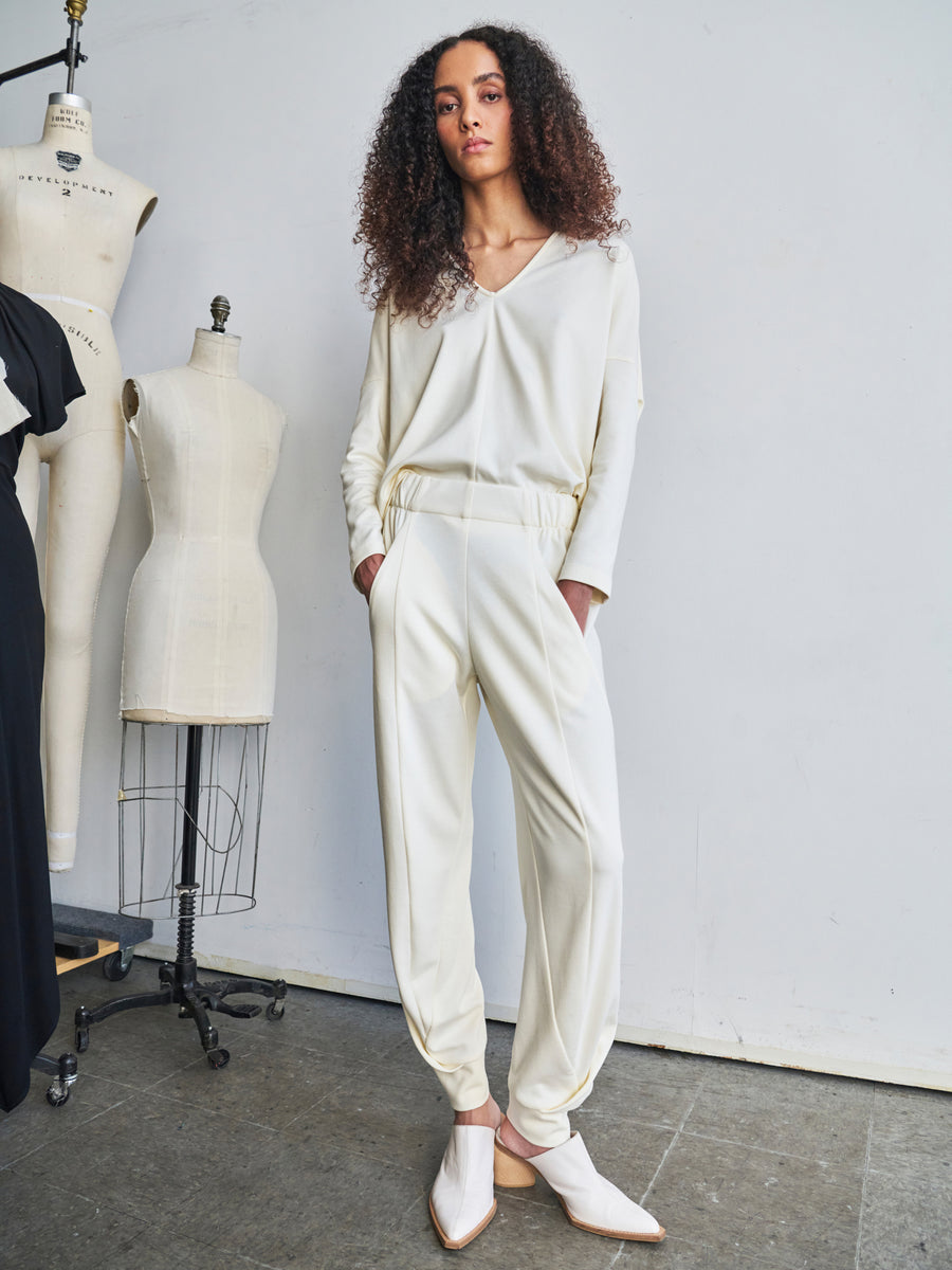 A person with curly hair stands in a studio, wearing a white long-sleeved top paired with the Jersey Cuff Akeo Pant from Zero + Maria Cornejo, both made from sustainable materials. Two dress forms are in the background on the concrete floor, and the wall is plain white.