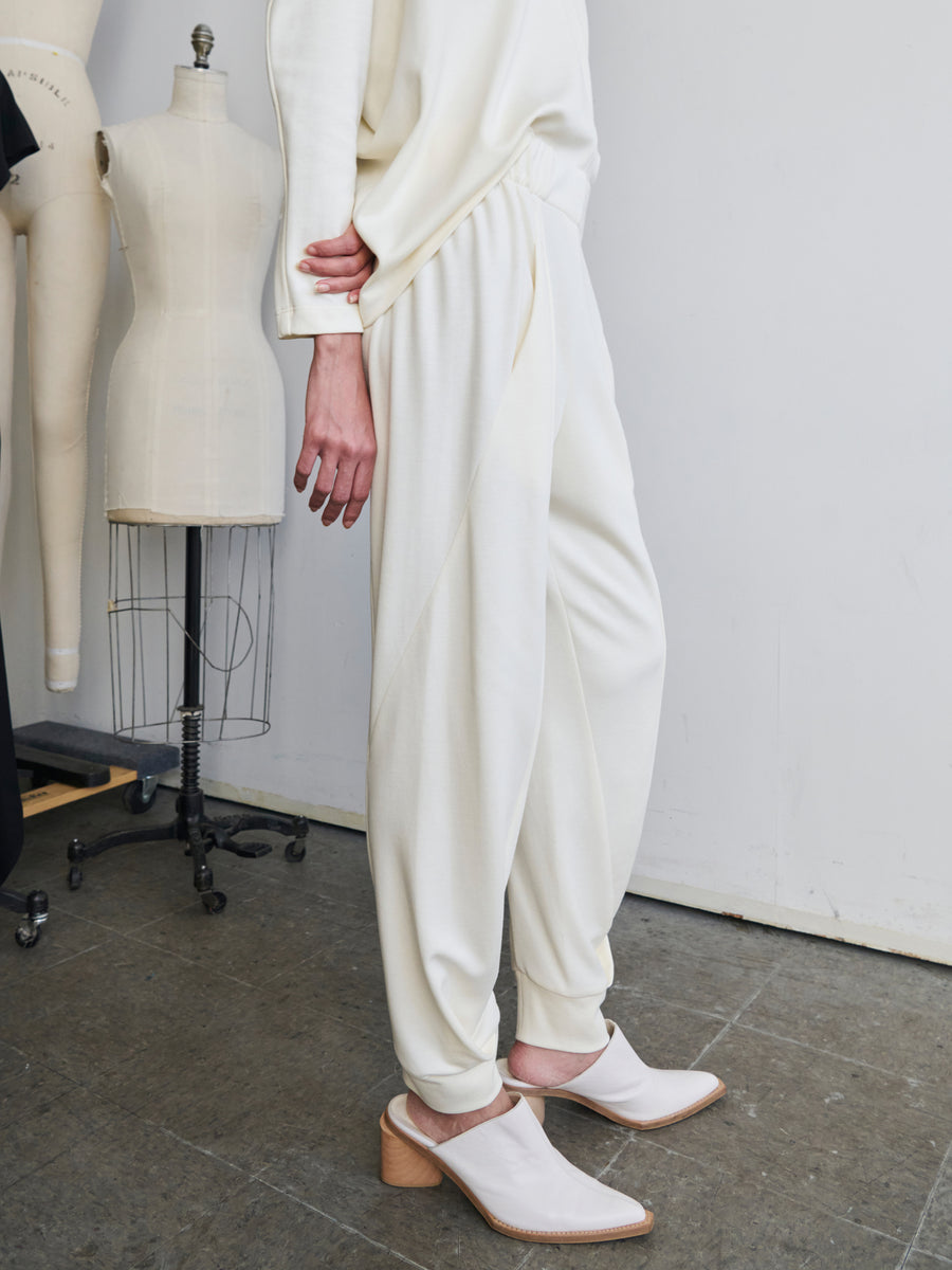 A person wearing a cream-colored outfit stands in a room with dress forms and sewing equipment. Dressed in Zero + Maria Cornejo's Jersey Cuff Akeo Pants paired with a matching long-sleeve top, crafted through sustainable production, they complete the look with white heeled clogs.