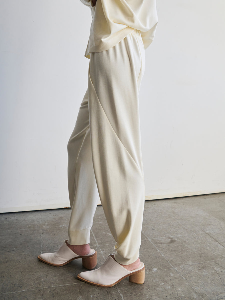 A person is standing on a concrete floor, dressed in cream-colored Zero + Maria Cornejo's Jersey Cuff Akeo Pant paired with a matching top. Their ensemble includes cream slip-on shoes with block heels. The outfit, crafted through sustainable production methods, is set against a plain white wall.