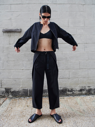 A person wearing black Zero + Maria Cornejo Wrap Akeo Pant, a black crop top, a black jacket, and black slide sandals stands outdoors against a gray concrete wall. They are posing with arms slightly raised and slightly bent while wearing sunglasses. Their hair is pulled back.