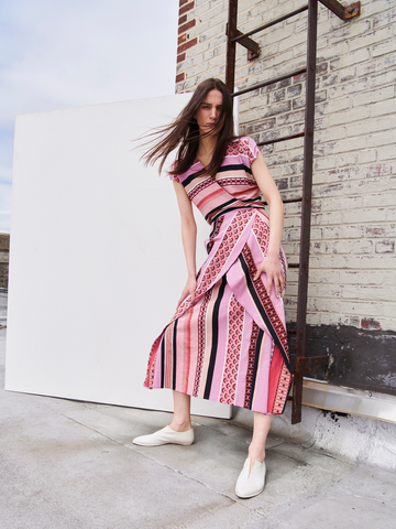 A person with long hair poses outdoors against a weathered brick wall and metal ladder, wearing the Mido Dress from Zero + Maria Cornejo. The pink and black striped jacquard wrap-effect v-neck dress and white shoes convey movement through their breezy stance.