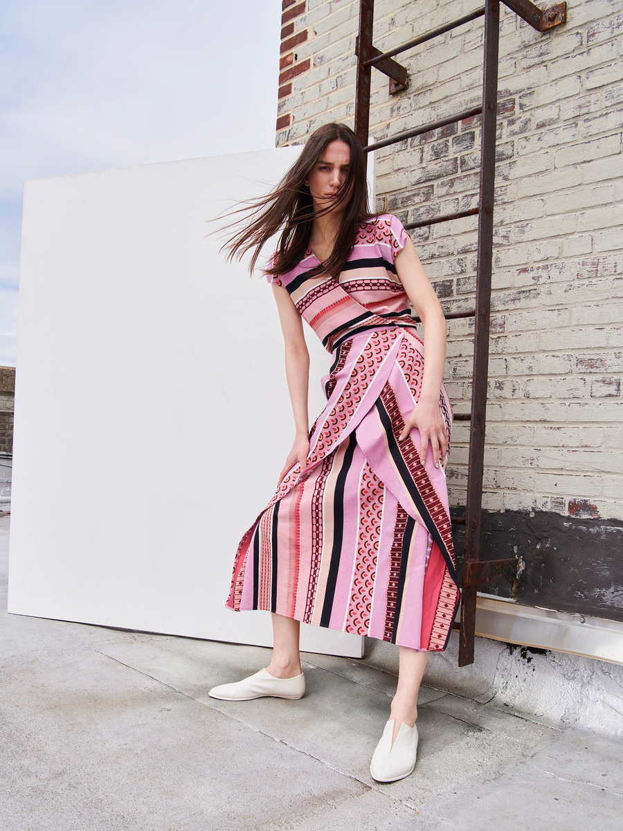 A person with long hair poses outdoors against a weathered brick wall and metal ladder, wearing the Mido Dress from Zero + Maria Cornejo. The pink and black striped jacquard wrap-effect v-neck dress and white shoes convey movement through their breezy stance.