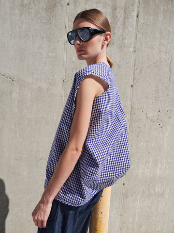 Wearing large black sunglasses and an Ina Tank by Zero + Maria Cornejo, which features a subtle houndstooth print and sleeveless v-neck design, a person stands against a concrete wall. With hair pulled back, they look to the side as sunlight casts intriguing shadows on the textured surface.