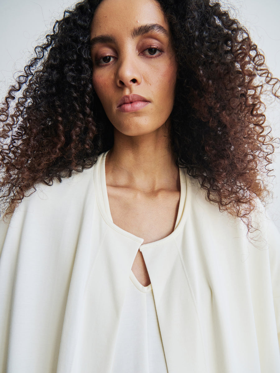 A woman with curly hair, dressed in a cream-colored Jersey Koya Coat by Zero + Maria Cornejo, gazes at the camera with a neutral expression against a plain, light background.