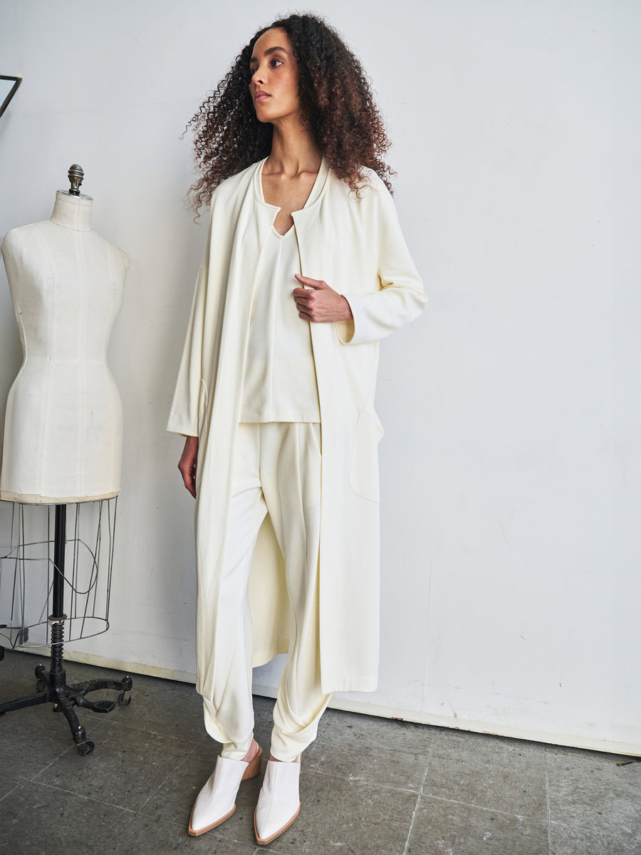 A person with curly hair is in a minimalistic studio, wearing the Jersey Koya Coat by Zero + Maria Cornejo. The coat, made of cotton-modal jersey, features a draped cocoon silhouette and is paired with ankle boots. A dress form stands beside them against a plain white wall.