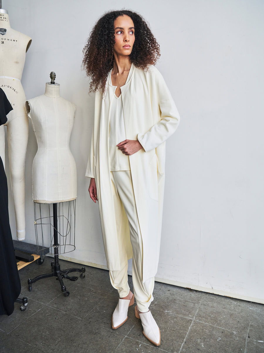 A person with curly hair stands on a concrete floor, wearing the Jersey Koya Coat from Zero + Maria Cornejo. The coat features a flowing off-white outfit with a draped cocoon silhouette and is paired with light-colored shoes. They are positioned next to two dress forms against a plain wall, embodying the elegance of bio-based materials.