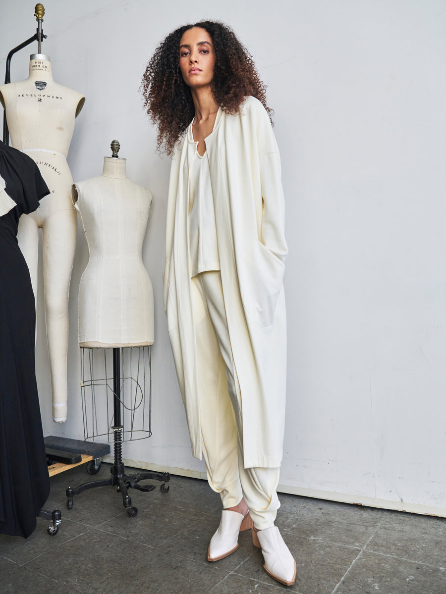 In a studio filled with dress forms, a person with curly hair stands confidently wearing the Jersey Koya Coat and loose pants by Zero + Maria Cornejo. The cream-colored cotton-modal ensemble exudes a fashionable and creative atmosphere with hints of draped cocoon silhouettes.