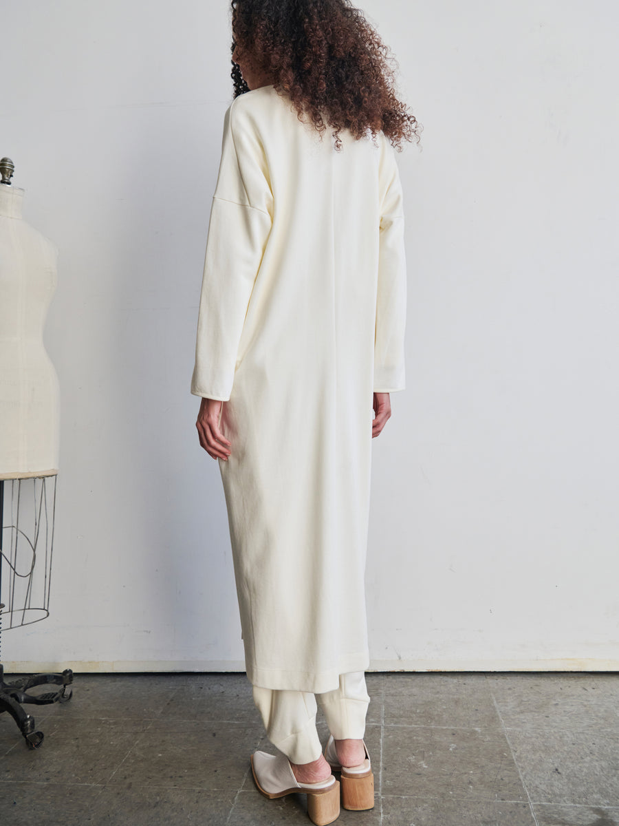 A person with curly hair is standing with their back to the camera, showcasing a long, light-colored Jersey Koya Coat and matching pants by Zero + Maria Cornejo. They are near a dress form in a minimalist setting with a concrete floor and plain wall, emphasizing the gentle drape of bio-based materials in a cocoon silhouette.