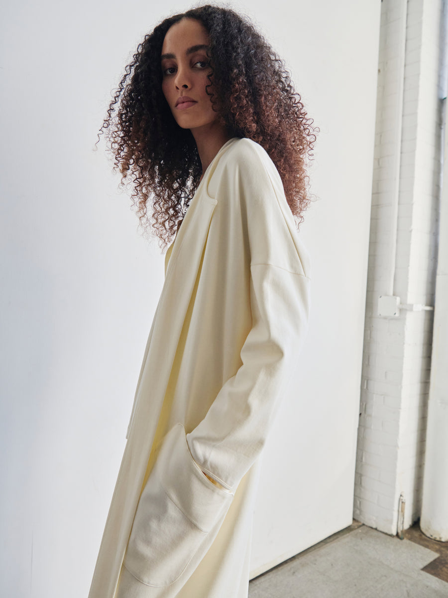 In a brightly lit room with a white brick wall, an individual with curly hair poses thoughtfully for the camera, elegantly wrapped in the draped cocoon silhouette of the Jersey Koya Coat by Zero + Maria Cornejo. This long, light-colored coat effortlessly combines elegance and comfort.