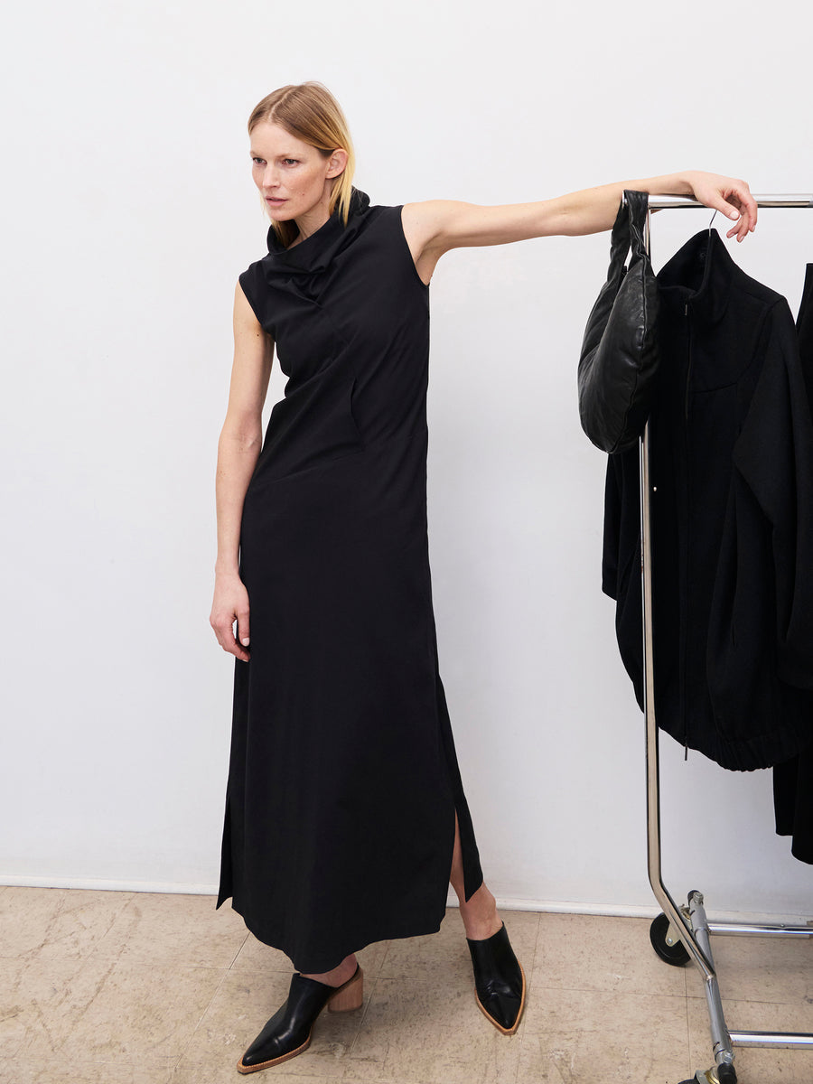 A person with short, light hair is posing indoors in a sleek, sleeveless Long Fold Dress by Zero + Maria Cornejo, featuring a high collar and side slits. They are leaning on a clothing rack with one arm, which has a black bag and black clothing hanging on it. The floor is a beige color.